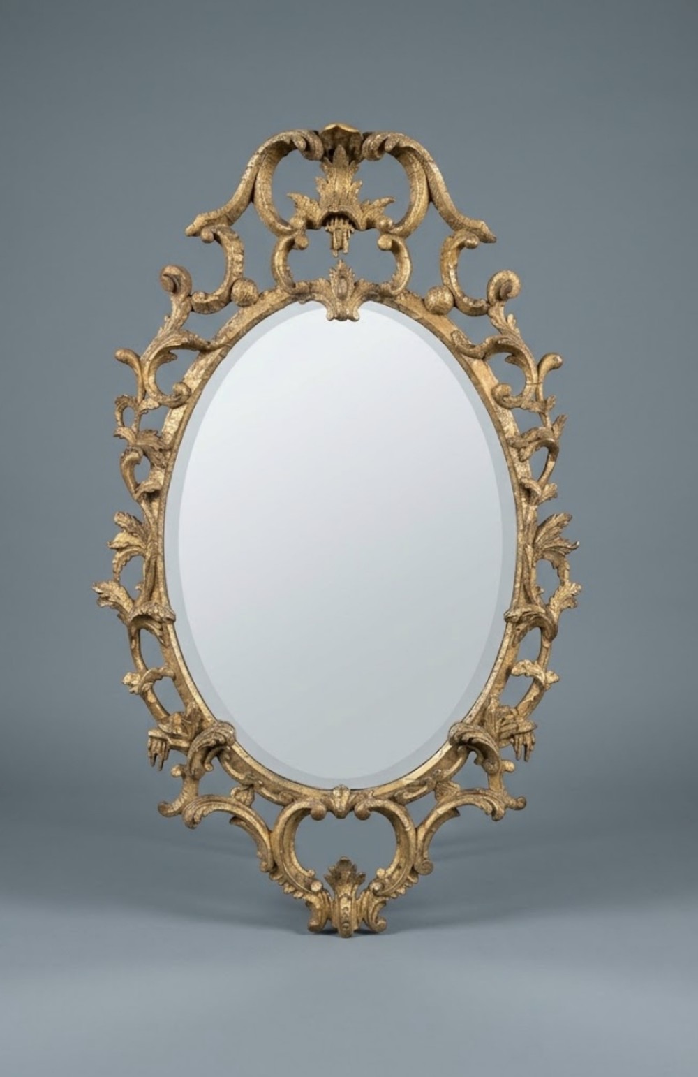 An 18th-century style oval wall mirror featuring a carved giltwood frame with openwork scrollwork and acanthus leaf cresting.