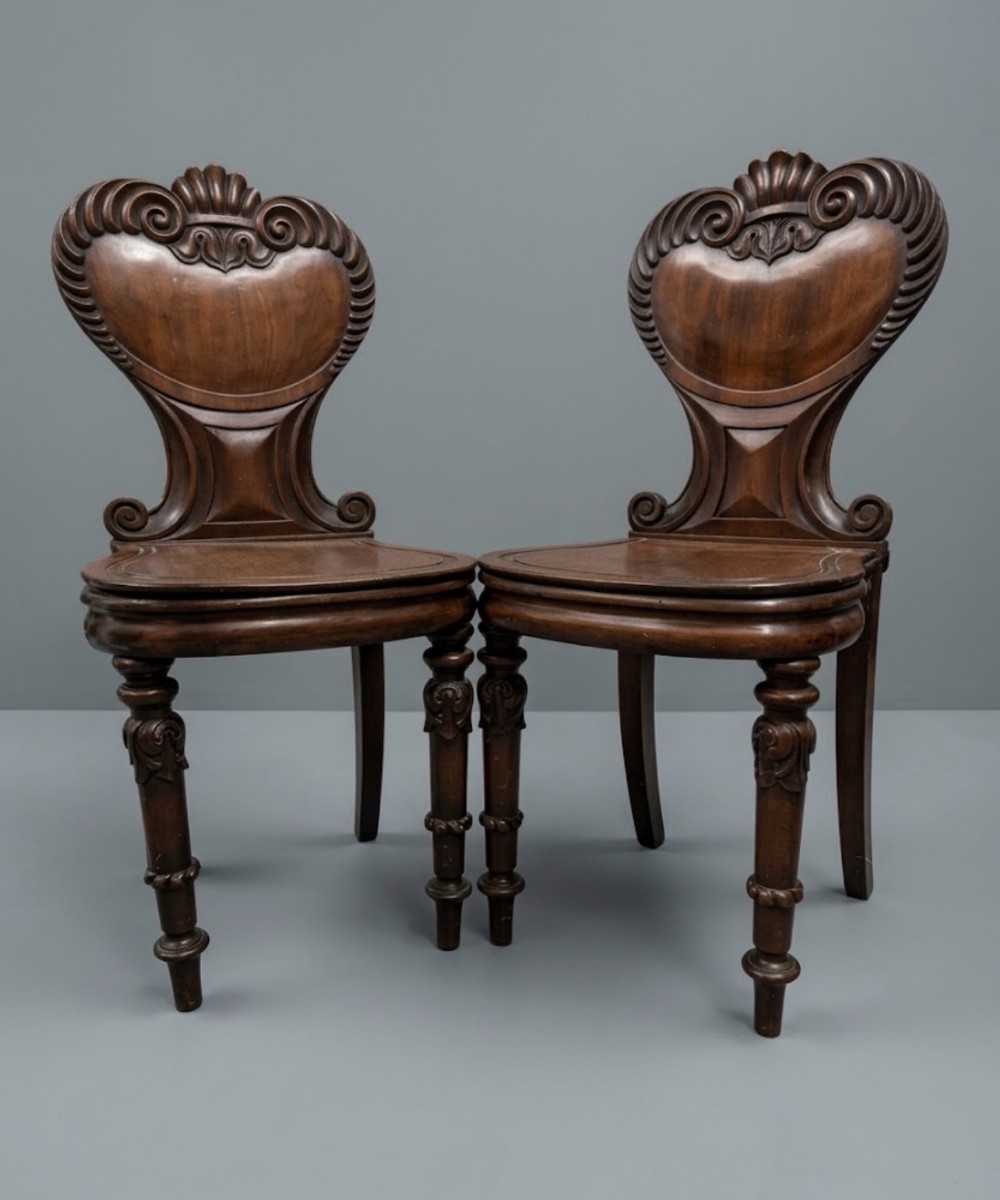 A pair of 19th-century carved mahogany hall chairs featuring intricate scroll cresting, solid timber seats, and turned front legs.