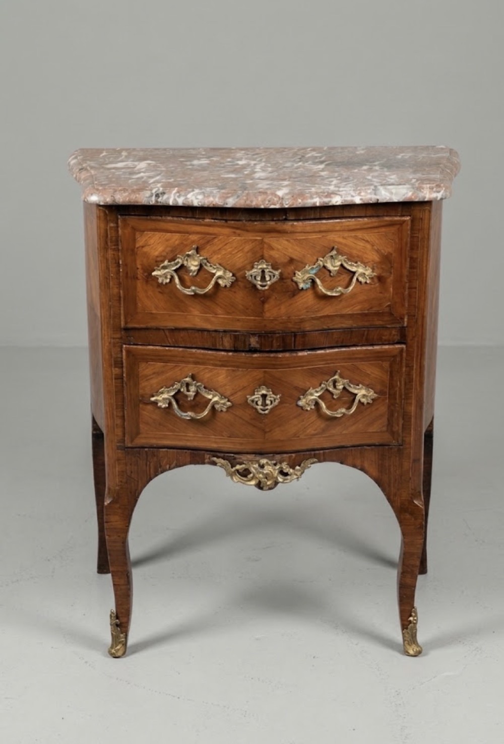 A 19th-century French two-drawer chest featuring a shaped pink marble top, geometric wood veneer inlay, curved cabriole legs, and decorative brass mounts.