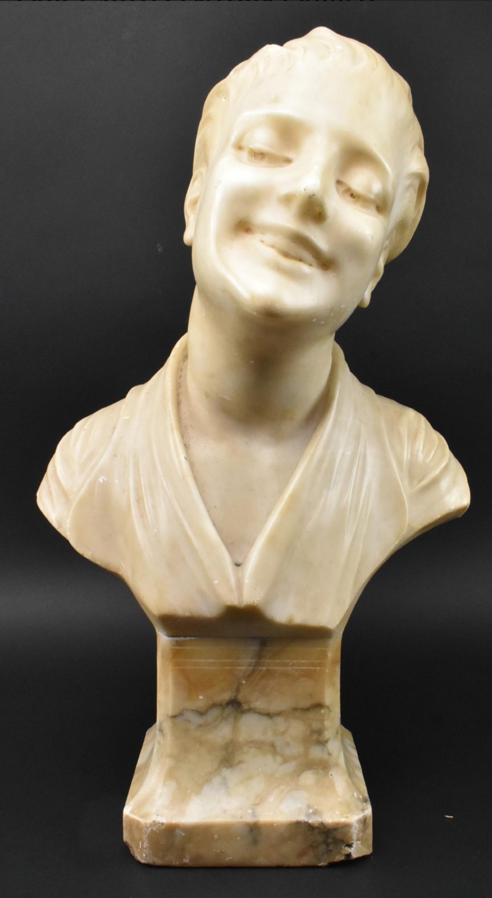 A carved cream alabaster bust depicting a smiling youth with a tilted head resting on an integral rectangular pedestal base.