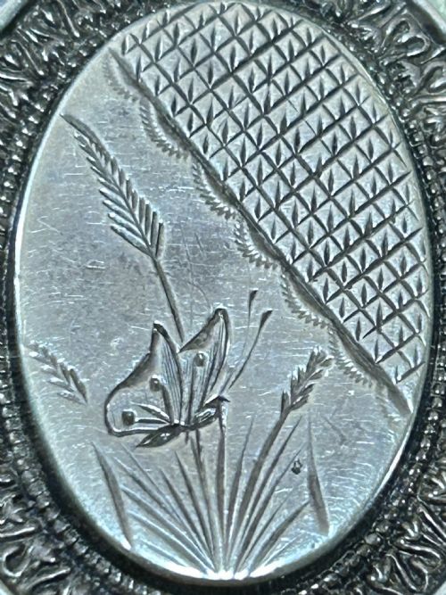 fig 1. An oval Victorian silver locket featuring engraved details of a butterfly, foliage, lattice textures, and decorative concentric borders.