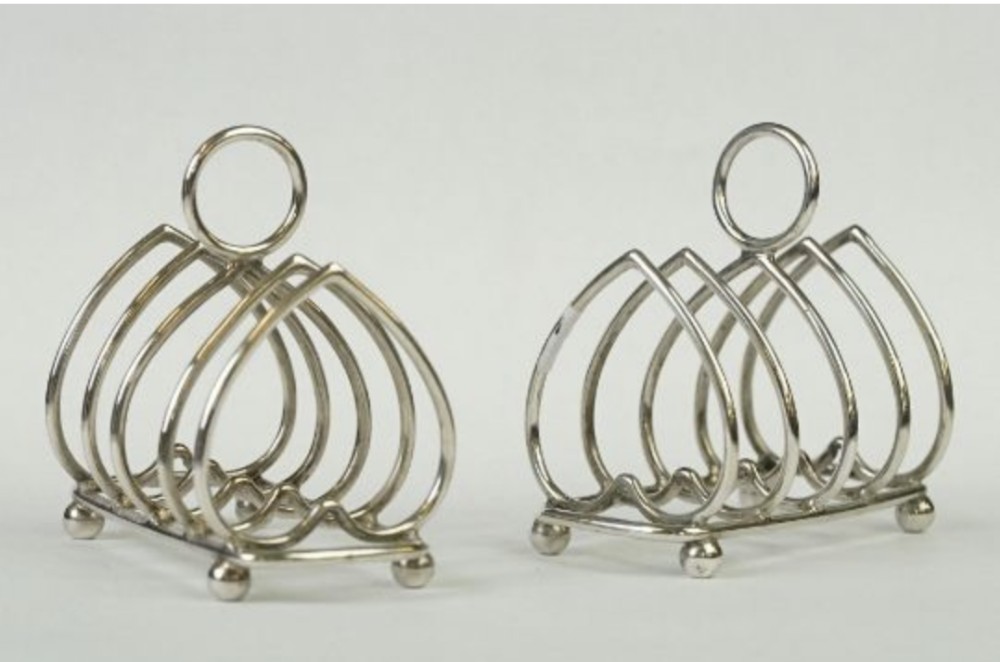 A pair of silver-toned metal toast racks featuring heart-shaped wire dividers, ball feet, and ring-shaped top handles.