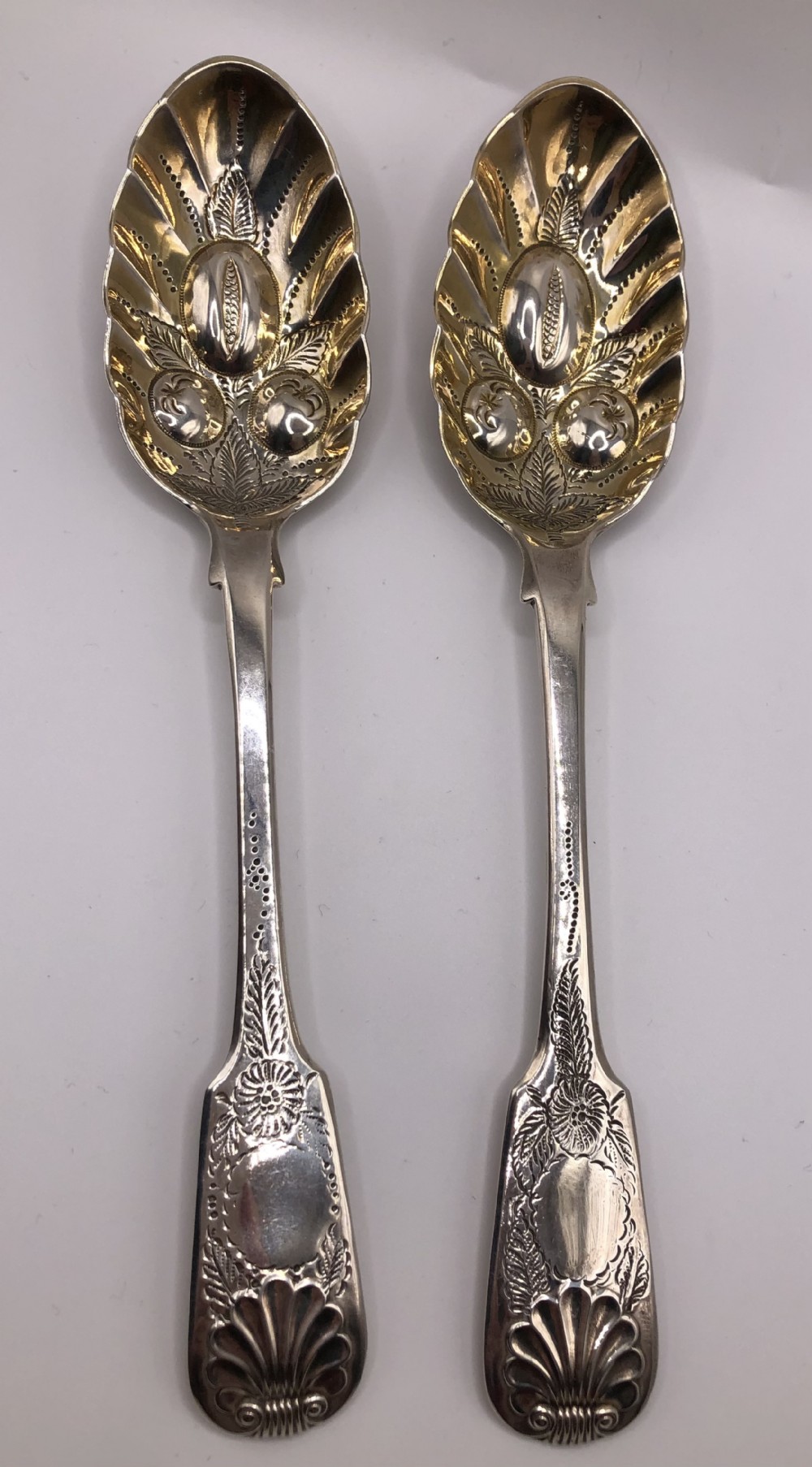 A pair of sterling silver serving spoons featuring gilded bowls with raised fruit motifs and elaborately chased handles ending in shell designs.