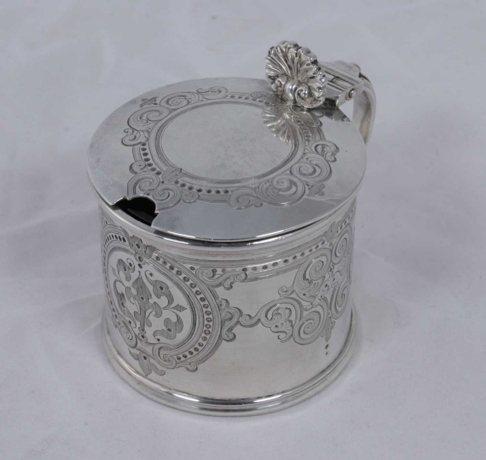 A cylindrical Victorian silver mustard pot featuring engraved scrollwork, a hinged lid with a shell thumbpiece, and a spoon cutout revealing a dark liner.