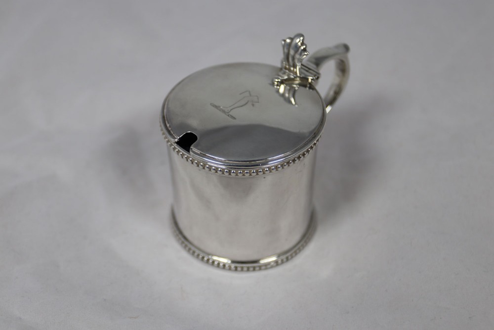 A cylindrical silver mustard pot featuring a hinged lid with a crest engraving, a shell thumbpiece, a side handle, and beaded trim around the top and base.