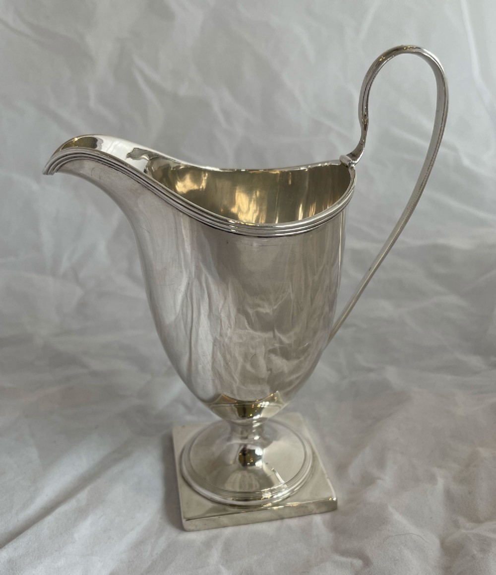 A polished silver helmet-shaped milk jug featuring a high loop handle and a pedestal stem on a square base.