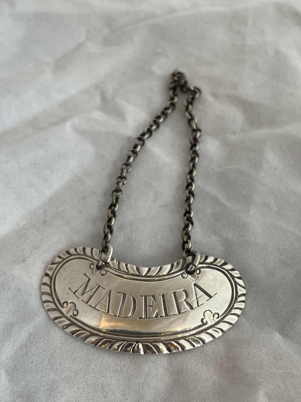 A crescent-shaped sterling silver decanter label engraved with Madeira and featuring a fluted border suspended on a chain.