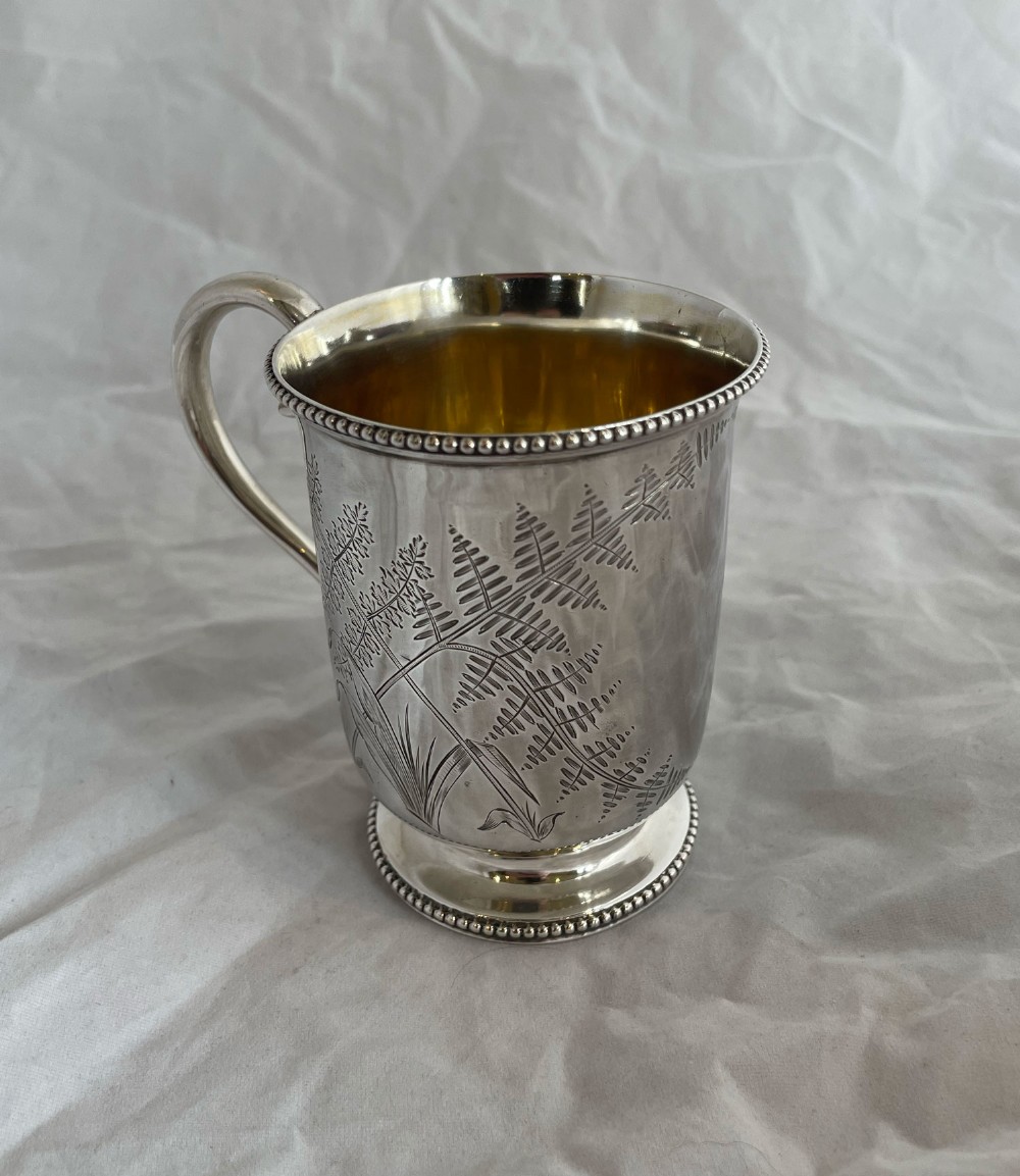 A Victorian silver christening mug featuring hand-engraved fern leaf motifs, a beaded rim border, and a gold-gilded interior.
