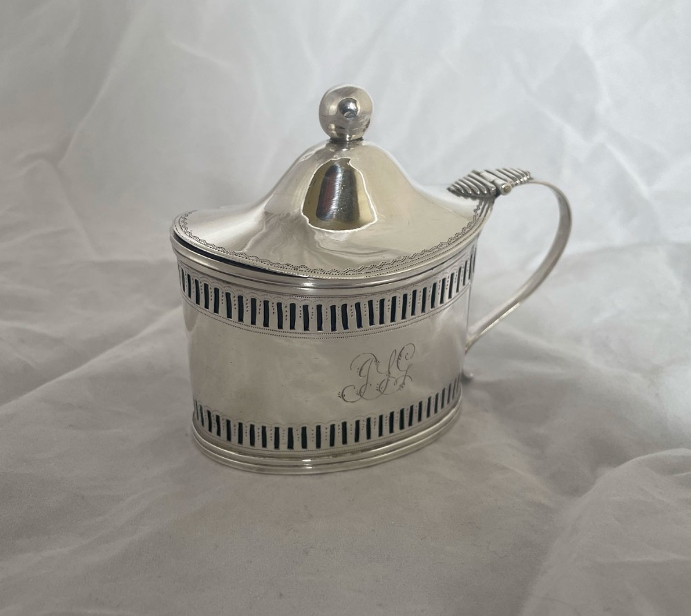 An oval George III sterling silver mustard pot featuring pierced geometric decoration, a hinged domed lid with finial, a scroll handle, and an engraved script monogram.