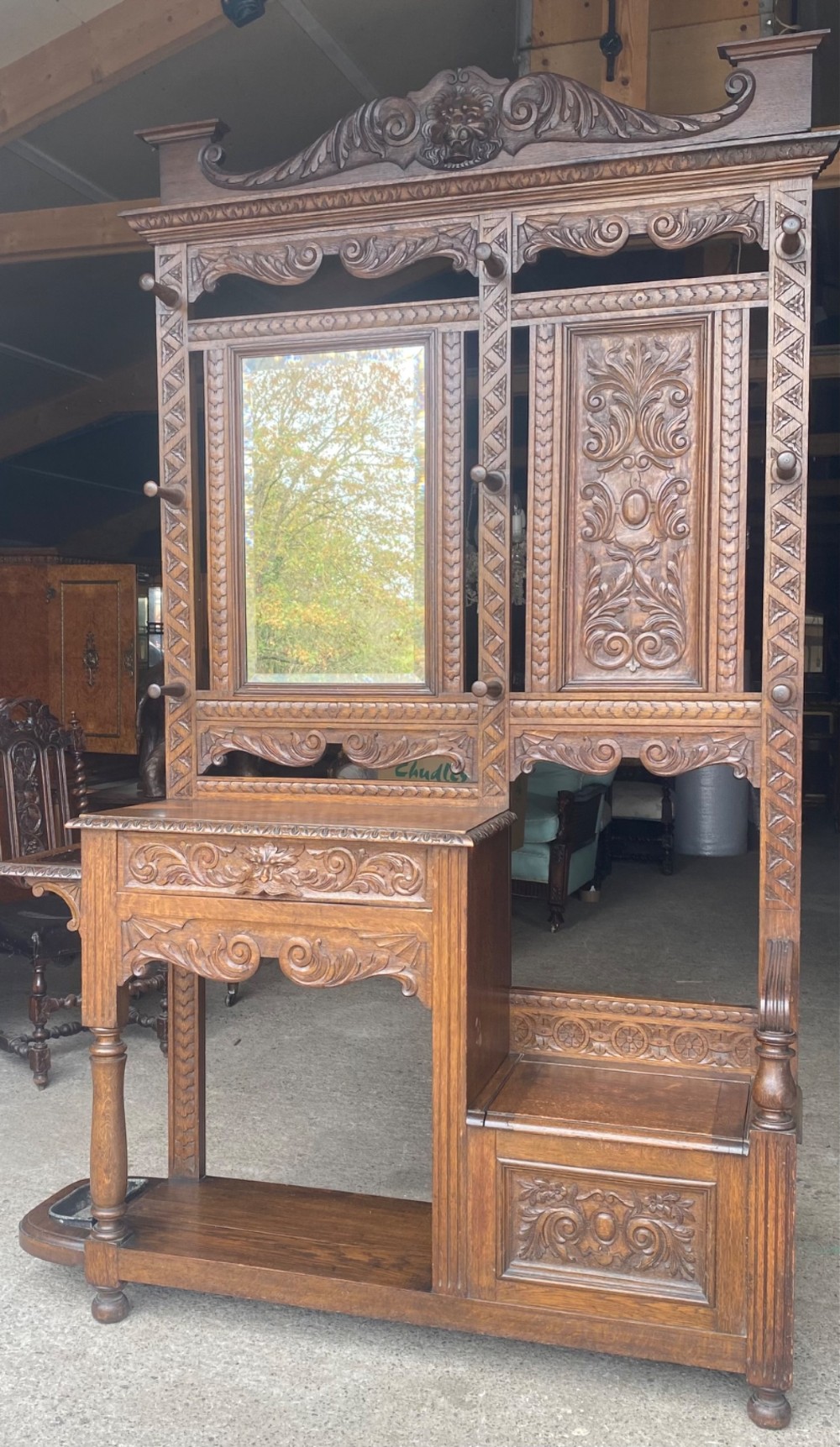 A tall carved oak hall stand featuring a central mirror, coat pegs, carved decorative panels, a small table section, and an umbrella holder with a drip tray.