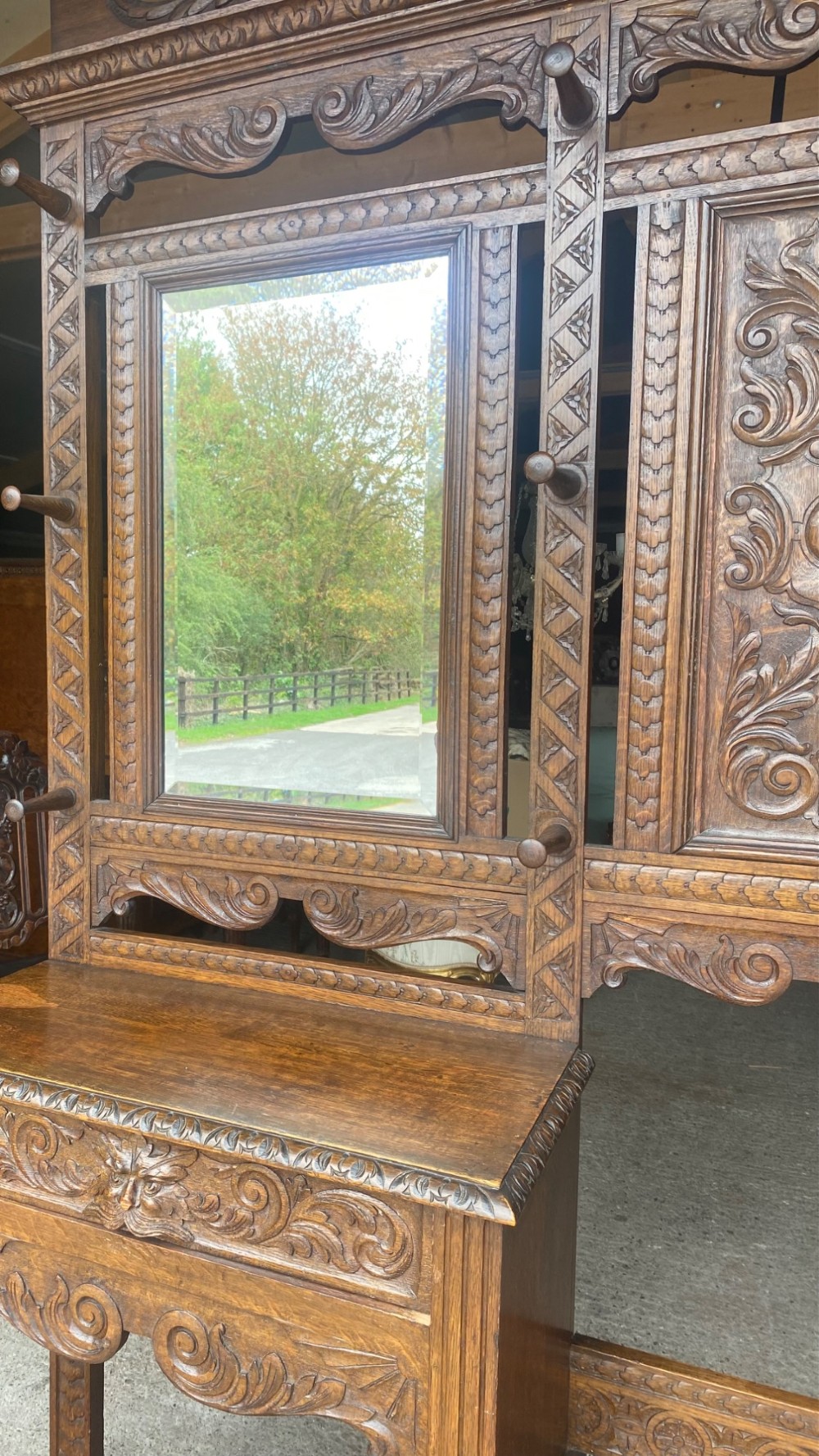 A tall carved oak hall stand featuring a central mirror, coat pegs, carved decorative panels, a small table section, and an umbrella holder with a drip tray. View II