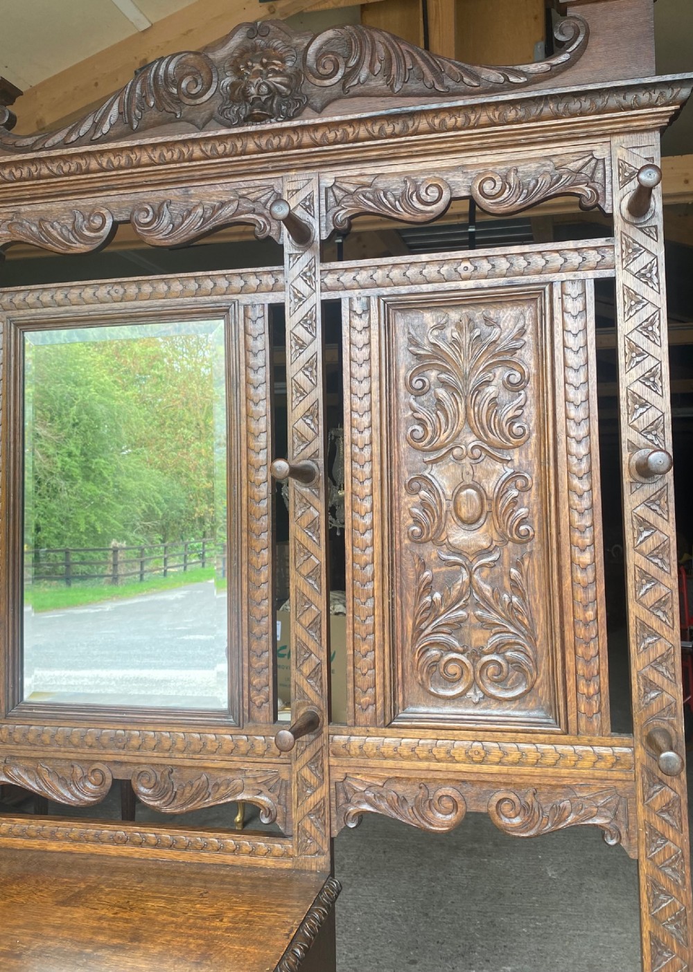A tall carved oak hall stand featuring a central mirror, coat pegs, carved decorative panels, a small table section, and an umbrella holder with a drip tray. View III