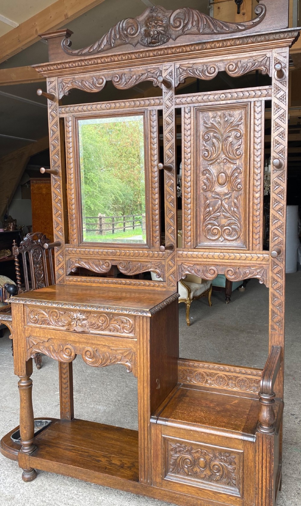 A tall carved oak hall stand featuring a central mirror, coat pegs, carved decorative panels, a small table section, and an umbrella holder with a drip tray. View IV