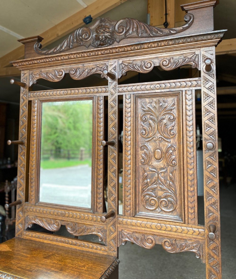A tall carved oak hall stand featuring a central mirror, coat pegs, carved decorative panels, a small table section, and an umbrella holder with a drip tray. View V