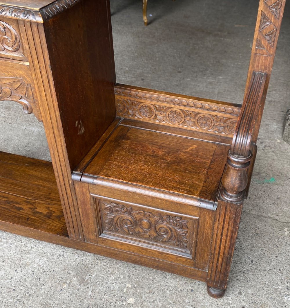 A tall carved oak hall stand featuring a central mirror, coat pegs, carved decorative panels, a small table section, and an umbrella holder with a drip tray. View VI