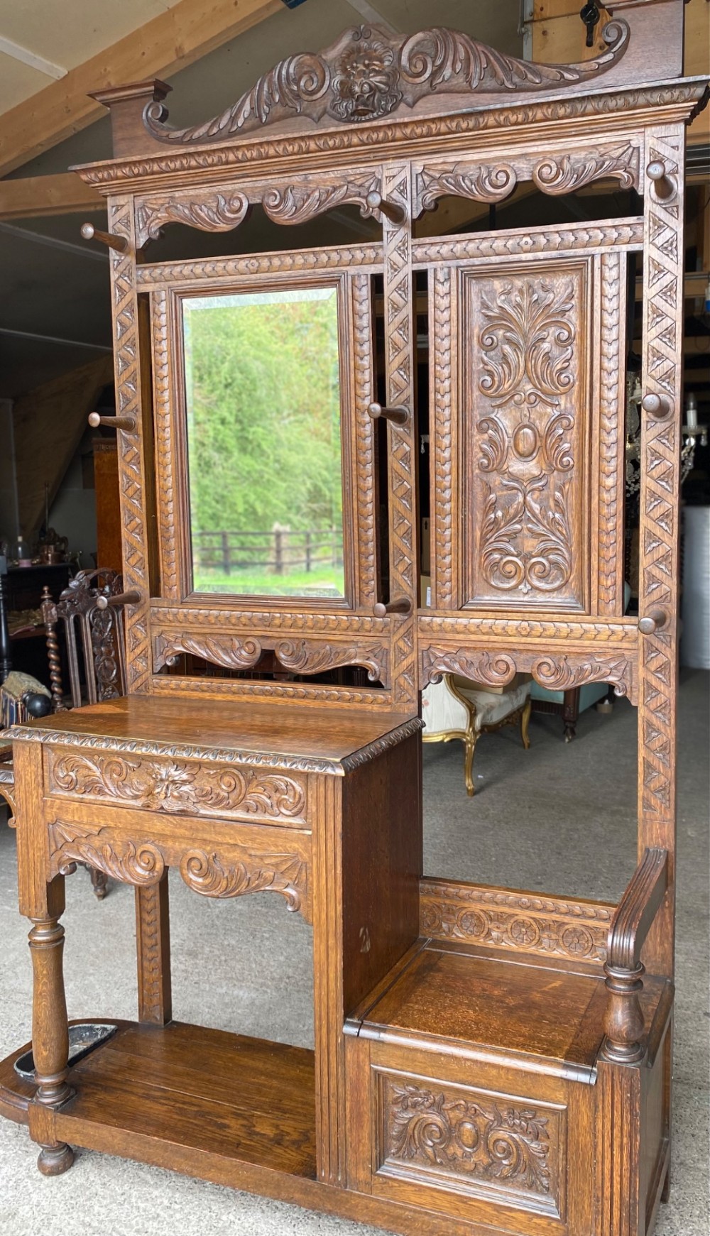 A tall carved oak hall stand featuring a central mirror, coat pegs, carved decorative panels, a small table section, and an umbrella holder with a drip tray. View VII