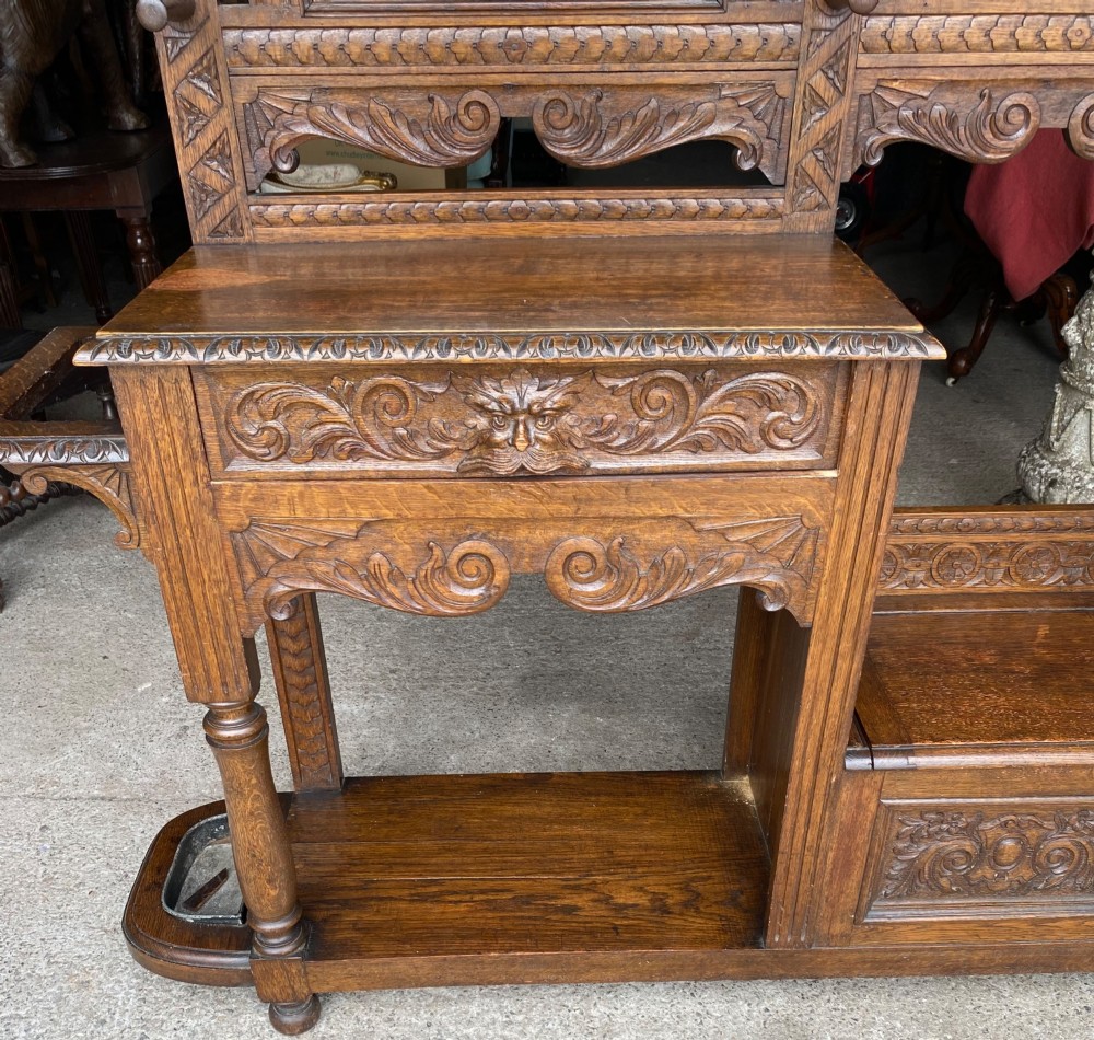 A tall carved oak hall stand featuring a central mirror, coat pegs, carved decorative panels, a small table section, and an umbrella holder with a drip tray. View VIII