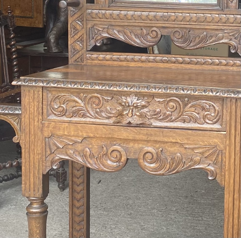 A tall carved oak hall stand featuring a central mirror, coat pegs, carved decorative panels, a small table section, and an umbrella holder with a drip tray. View X