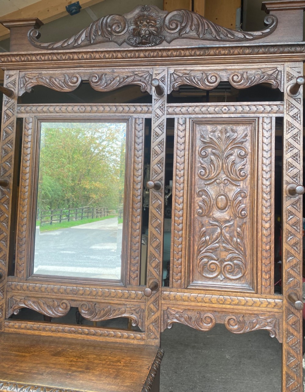 A tall carved oak hall stand featuring a central mirror, coat pegs, carved decorative panels, a small table section, and an umbrella holder with a drip tray. View XI