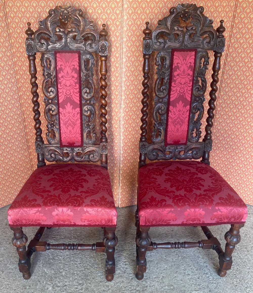 A pair of high-backed carved oak hall chairs featuring elaborate scrollwork, barley twist pillars, and red pattern damask upholstery.