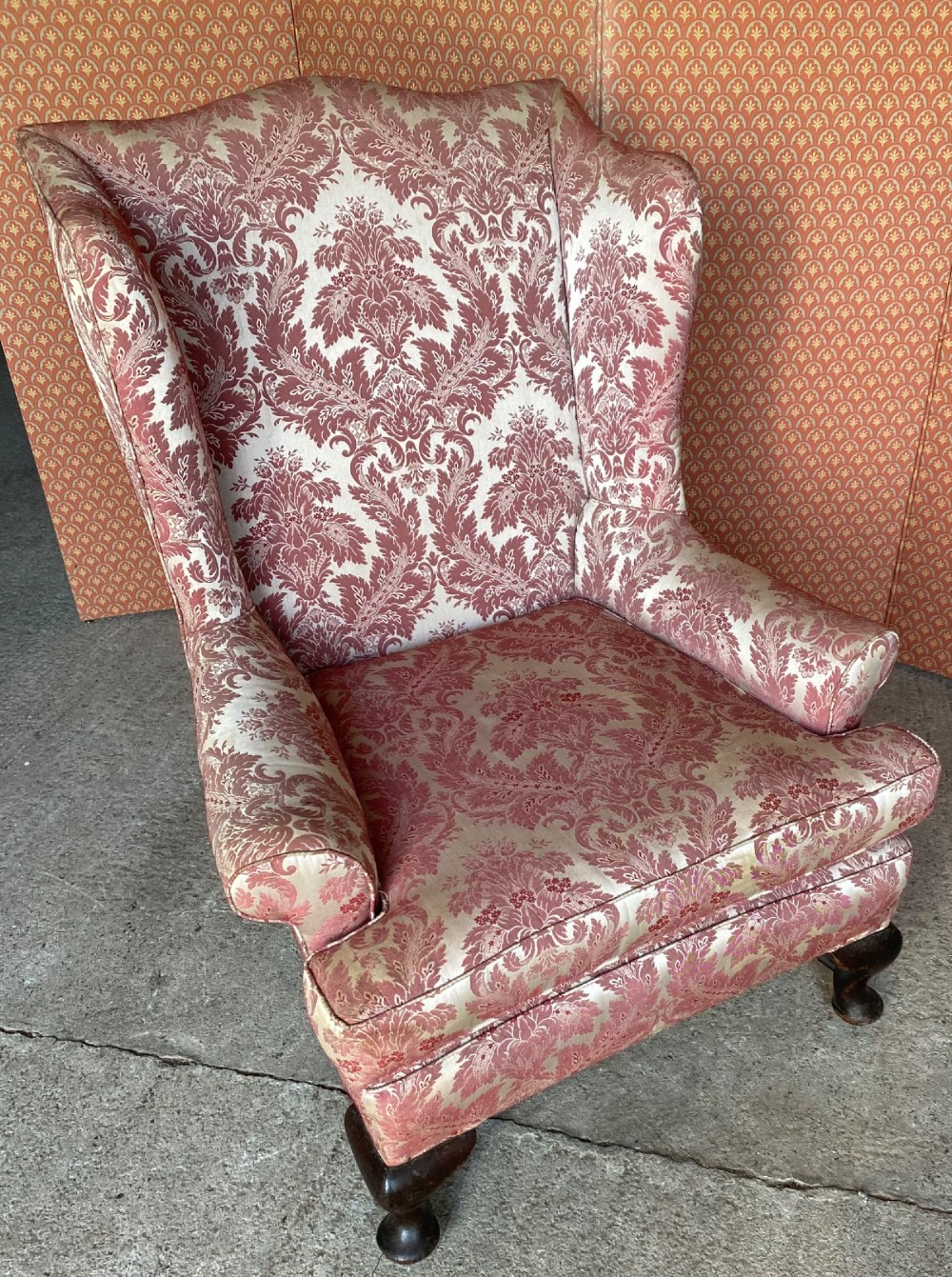 A high-back wingback armchair upholstered in rose pink and cream damask fabric, featuring rolled arms, a fitted seat cushion, and dark carved wooden legs. View V