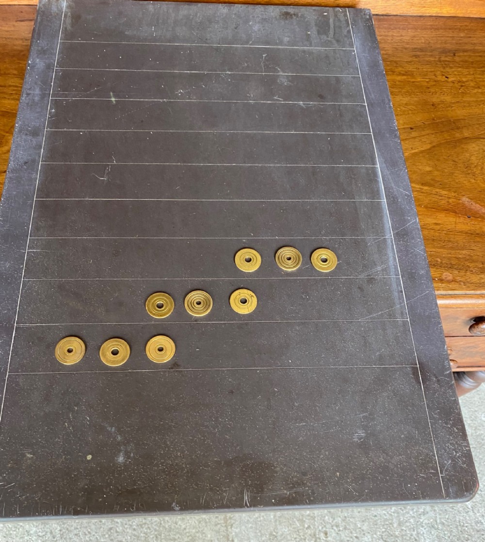 A rectangular dark grey slate shove hapenny board incised with horizontal scoring lines and several brass tokens.
