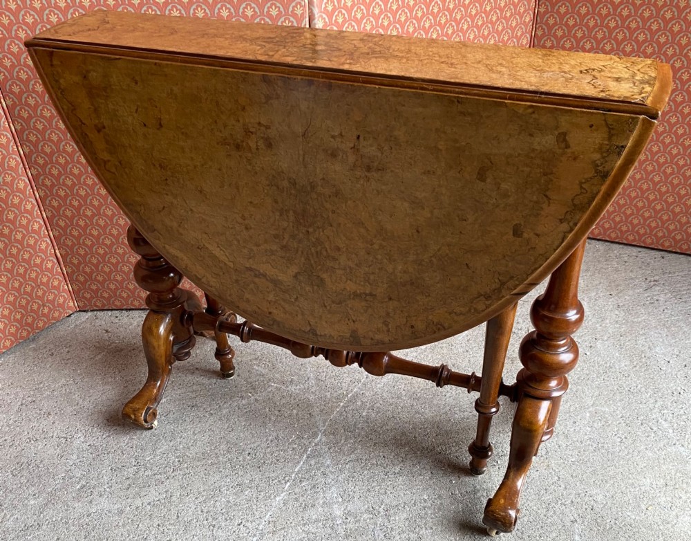 A Victorian burr walnut drop leaf table with turned twin column supports, a stretchered base, scrolled feet on brass castors, and a figured veneer surface.