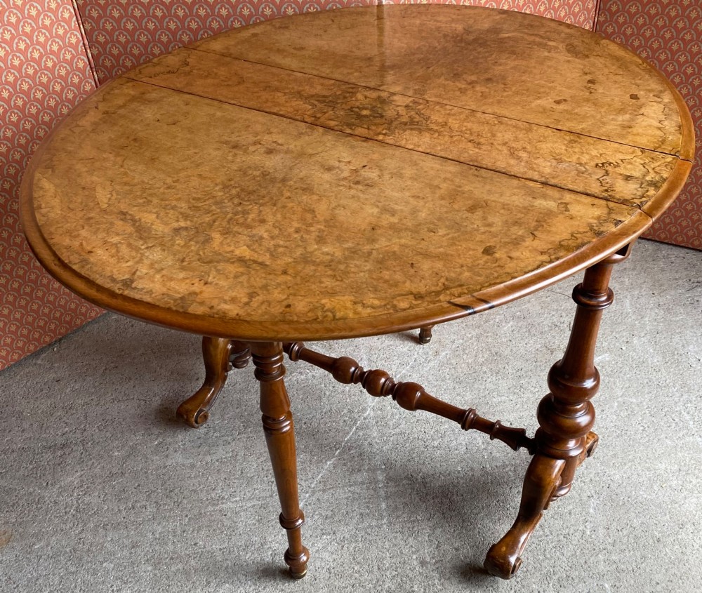 A Victorian burr walnut drop leaf table with turned twin column supports, a stretchered base, scrolled feet on brass castors, and a figured veneer surface. View VI