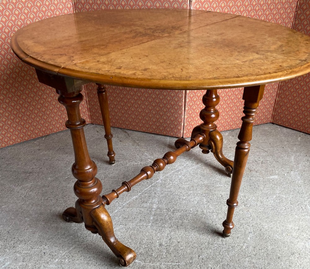 A Victorian burr walnut drop leaf table with turned twin column supports, a stretchered base, scrolled feet on brass castors, and a figured veneer surface. View VII