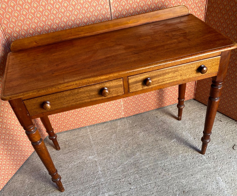 A Victorian warm brown mahogany writing desk featuring two frieze drawers with turned wooden knobs, supported by turned tapered legs and a low gallery backboard. View VI