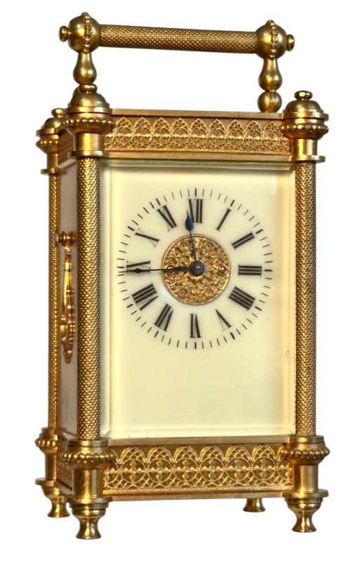 fig 1. An antique French gilt brass carriage clock featuring detailed filigree bands, turned corner columns, a white enamel dial with Roman numerals, and a top carrying handle.