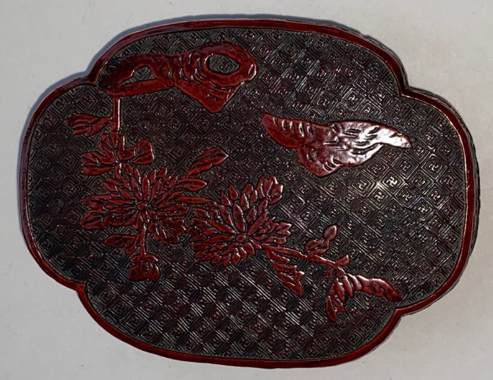 A lobed Qing Dynasty carved red cinnabar lacquer lidded box decorated with chrysanthemum blossoms and rockwork over a dark diaper background. View II