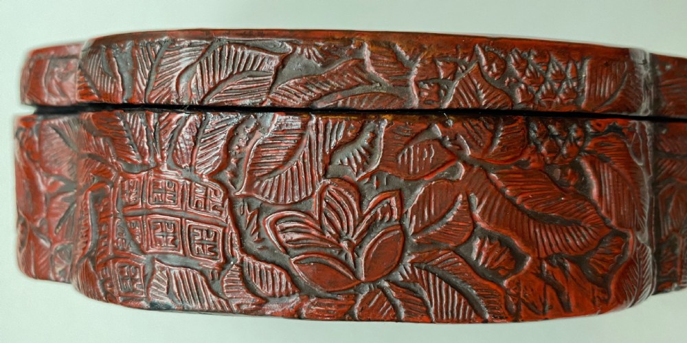 A lobed Qing Dynasty carved red cinnabar lacquer lidded box decorated with chrysanthemum blossoms and rockwork over a dark diaper background. View VI