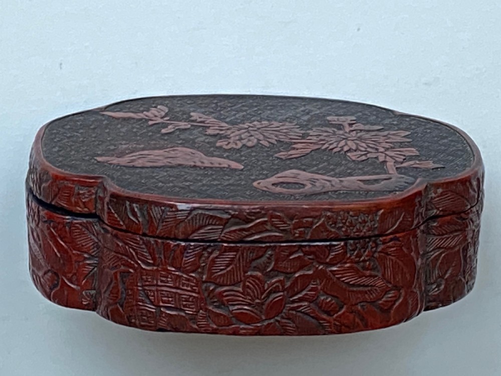 A lobed Qing Dynasty carved red cinnabar lacquer lidded box decorated with chrysanthemum blossoms and rockwork over a dark diaper background. View VII