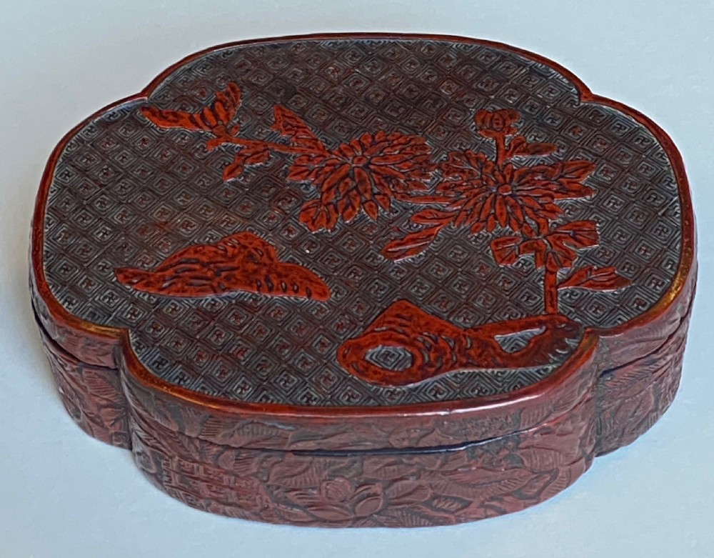 A lobed Qing Dynasty carved red cinnabar lacquer lidded box decorated with chrysanthemum blossoms and rockwork over a dark diaper background. View X