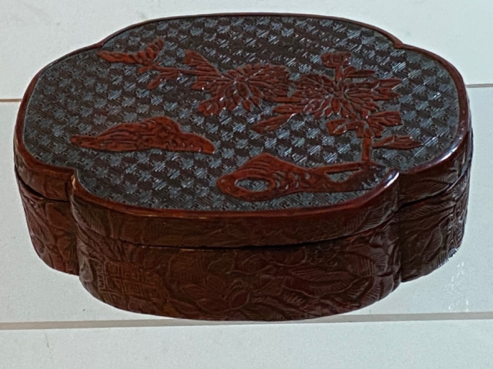 A lobed Qing Dynasty carved red cinnabar lacquer lidded box decorated with chrysanthemum blossoms and rockwork over a dark diaper background. View XII