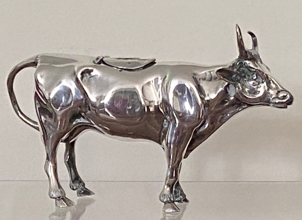 A sculpted silver cow creamer featuring a hollow body, looped tail handle, curved horns, and a hinged flap on its back. View II