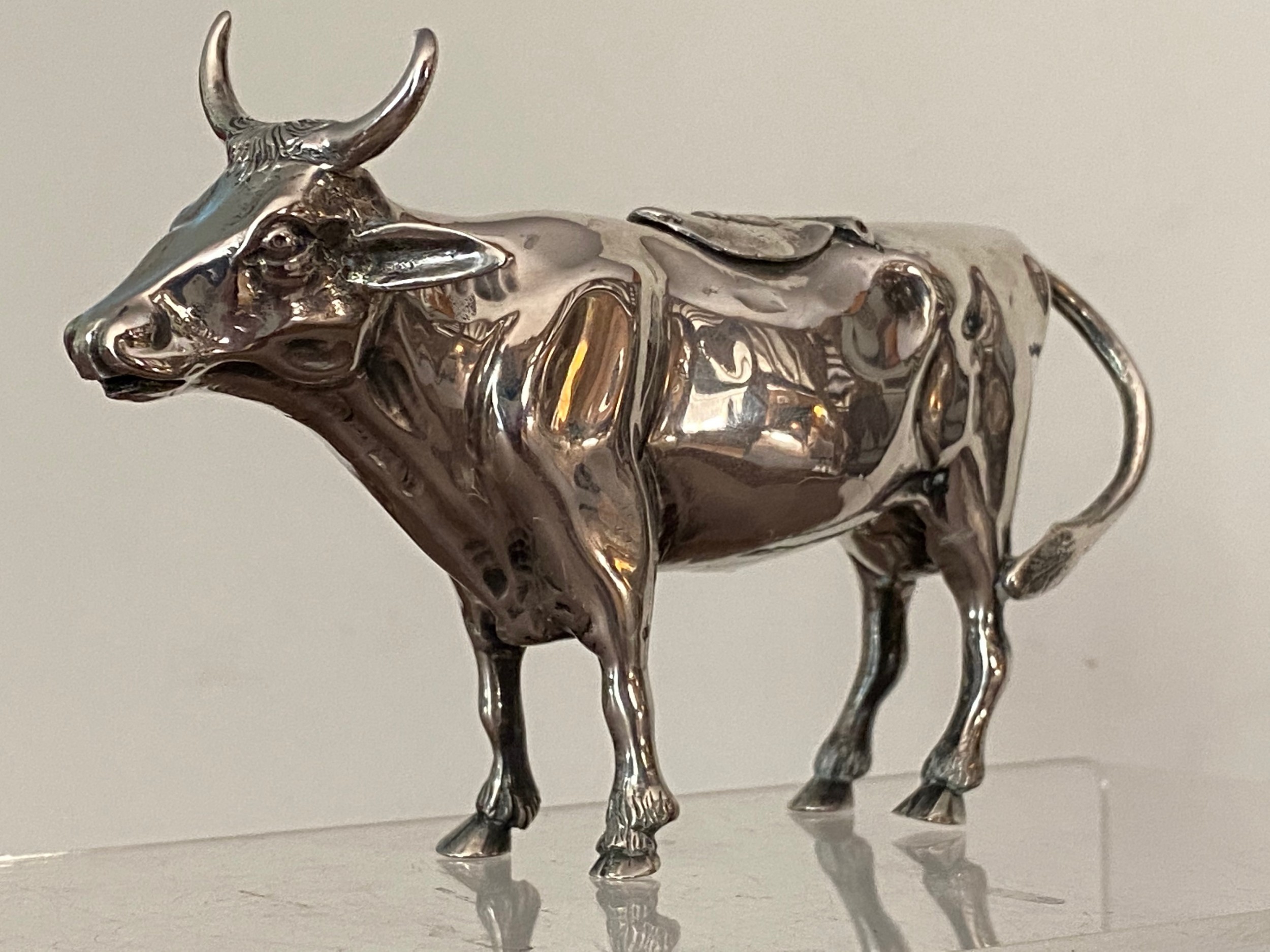 A sculpted silver cow creamer featuring a hollow body, looped tail handle, curved horns, and a hinged flap on its back.