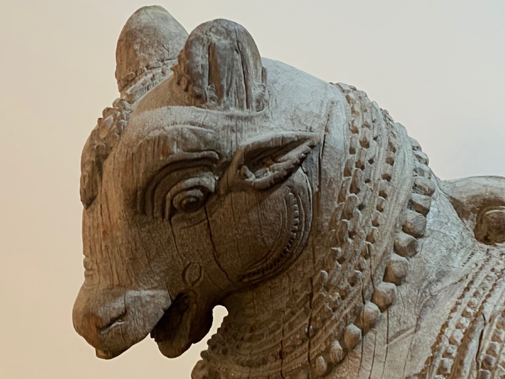 A carved wooden sculpture of a seated Nandi bull on a decorated plinth featuring intricate neck garlands and a saddle cover. View VI