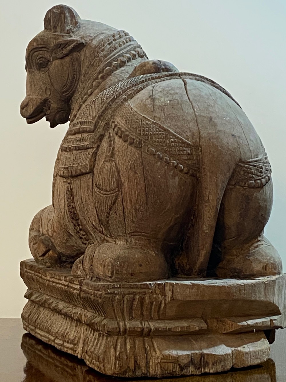 A carved wooden sculpture of a seated Nandi bull on a decorated plinth featuring intricate neck garlands and a saddle cover. View VII