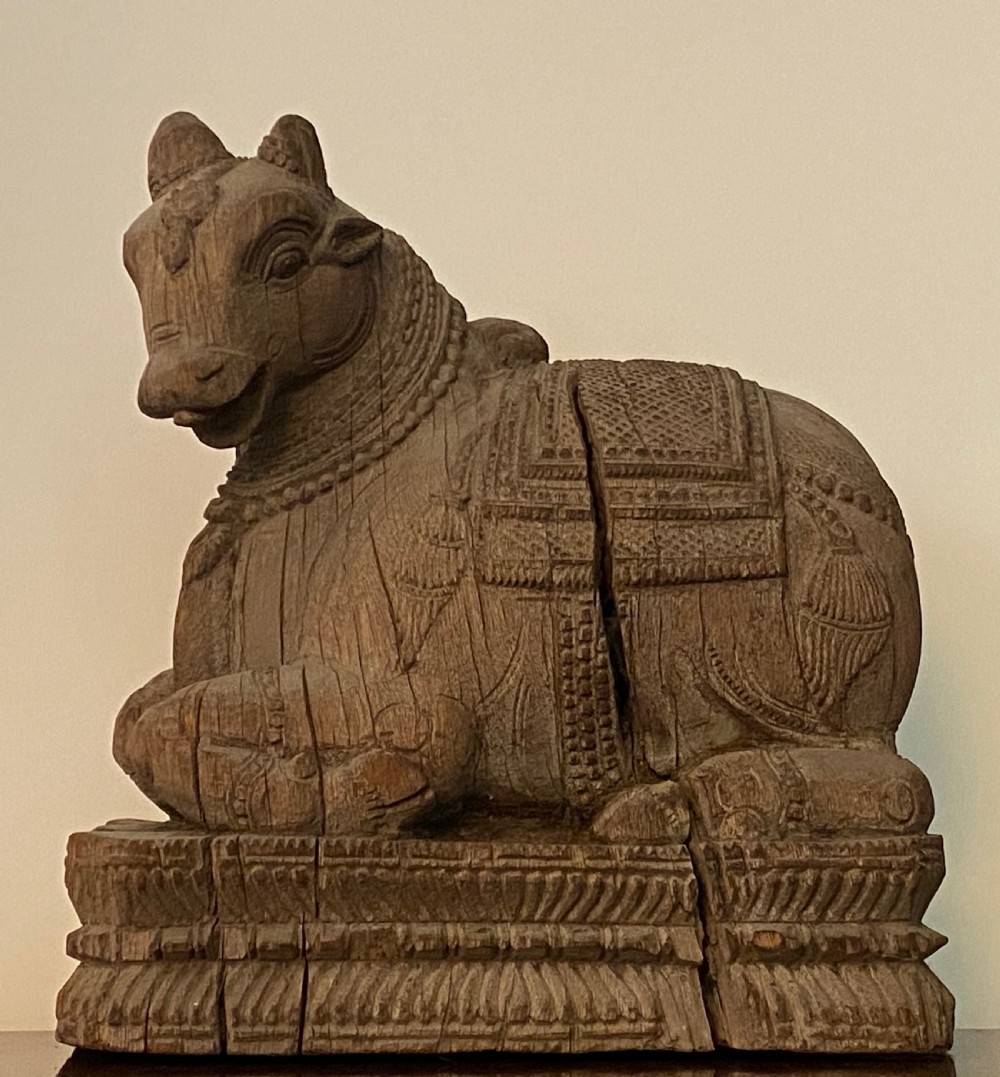 A carved wooden sculpture of a seated Nandi bull on a decorated plinth featuring intricate neck garlands and a saddle cover. View X