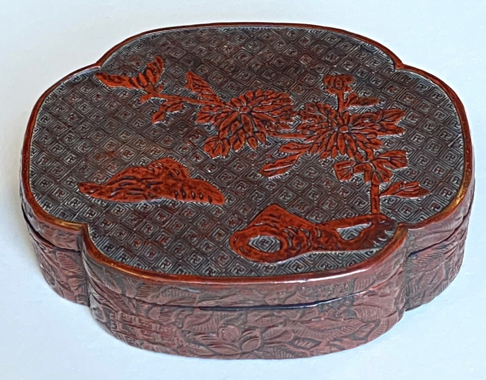 A lobed Qing Dynasty carved red cinnabar lacquer lidded box decorated with chrysanthemum blossoms and rockwork over a dark diaper background.