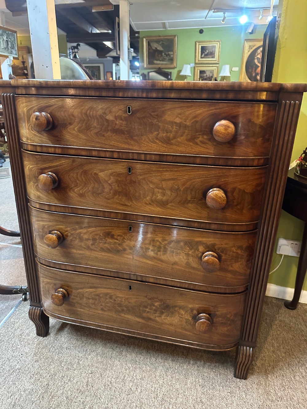 A Regency flame mahogany chest of drawers featuring four graduated bow-front drawers, turned wood handles, and reeded side columns. View X