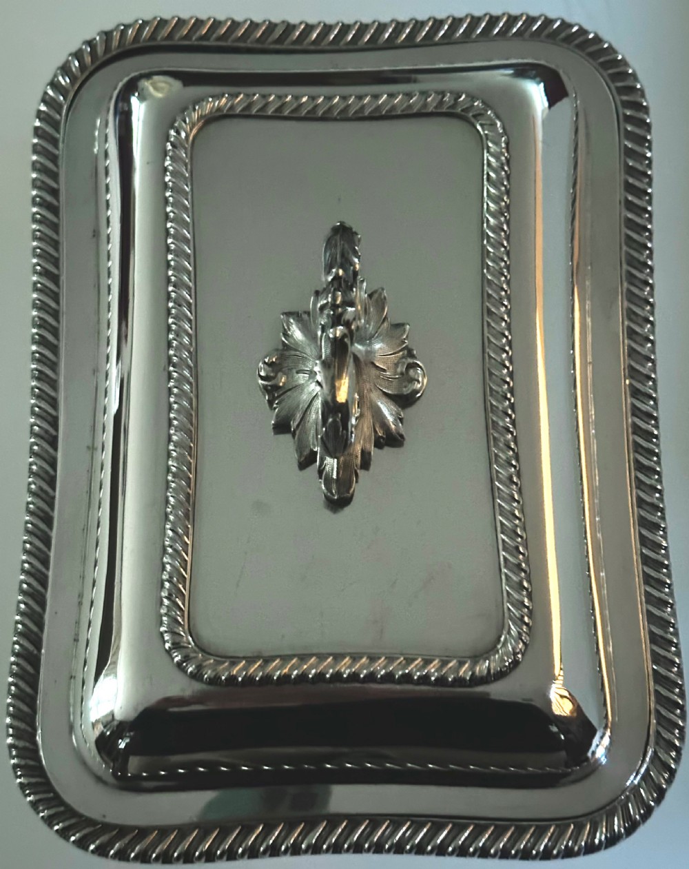 A rectangular silver-plated covered serving dish featuring gadrooned rope borders and a decorative foliage-detailed handle on top. View II