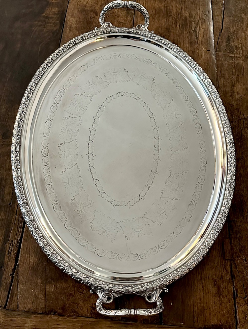 A large oval Edwardian silver-plated serving tray with detailed scrollwork rims, surface foliage engraving, and two cast side handles.