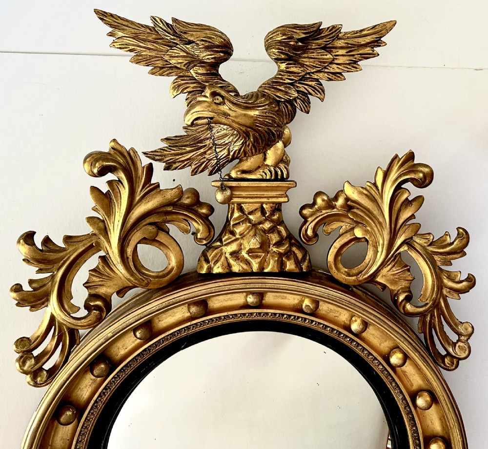 A circular Regency giltwood convex mirror featuring a carved eagle cresting, acanthus leaf scrolls, and ball mouldings around an ebonised inner slip. View II