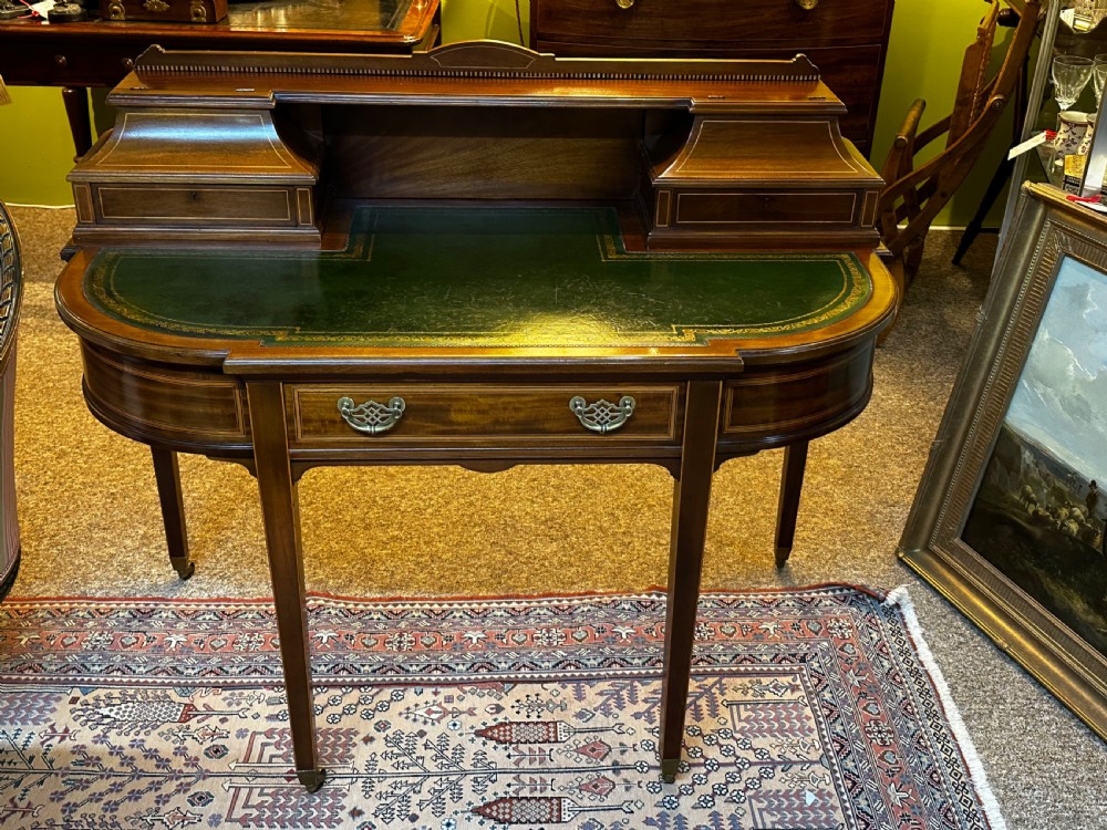 An antique mahogany writing desk featuring a green leather surface, a raised central superstructure with small drawers, satinwood string inlay, and tapered legs on brass castors.