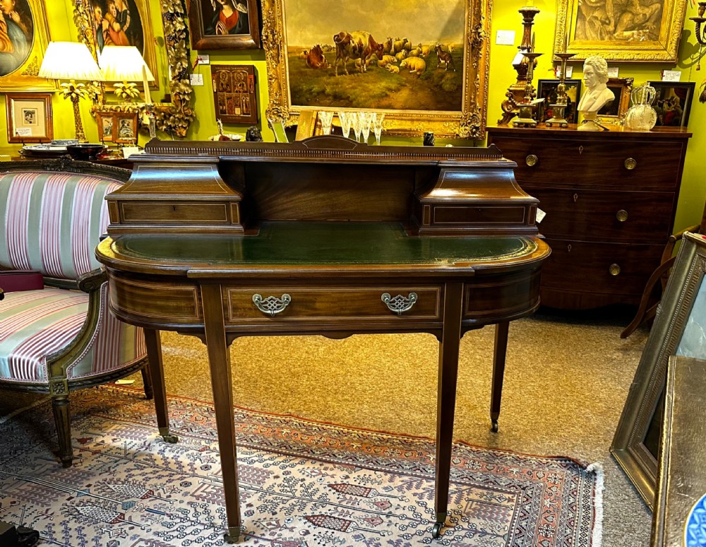 An antique mahogany writing desk featuring a green leather surface, a raised central superstructure with small drawers, satinwood string inlay, and tapered legs on brass castors. View III