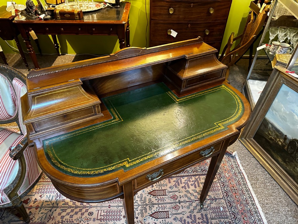 An antique mahogany writing desk featuring a green leather surface, a raised central superstructure with small drawers, satinwood string inlay, and tapered legs on brass castors. View IV