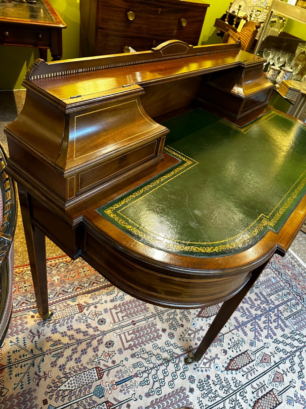 An antique mahogany writing desk featuring a green leather surface, a raised central superstructure with small drawers, satinwood string inlay, and tapered legs on brass castors. View V